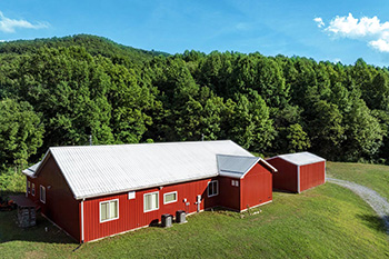 huge red metal barn with white roof and outbuildings sits on a grassy knoll in front of green mountain range