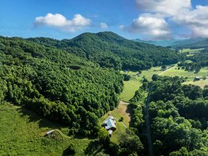 dramatic overhead shot of twisting valley surrounded by majestic, tree-covered mountains. In the foreground is the bright white roofline of a large barndominium and there are dramatic clouds in the sky
