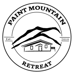 a black and white logo showing a drawing of a house in front of mountains and the words Paint Mountain Retreat in a circle