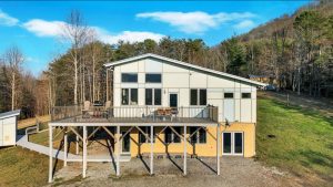 an angular contemporary house with a wall of windows, a huge deck and a lower level with glass french doors that open onto a walkway leading to a fancy shed
