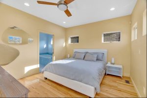 Sunlit primary bedroom with hardwood floors, ceiling fan and modern wall mirrors that reflect the view from the outside