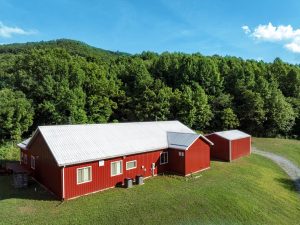 bright red metal barndominium sits on a green lawn with a forest behind it